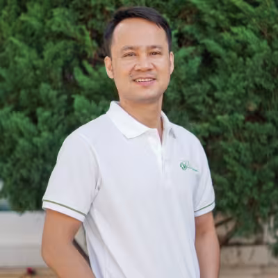 Phuc smiles warmly, wearing a white VVOB polo shirt. He stands against a backdrop of lush green foliage.
