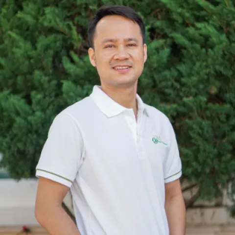 Phuc smiles warmly, wearing a white VVOB polo shirt. He stands against a backdrop of lush green foliage.