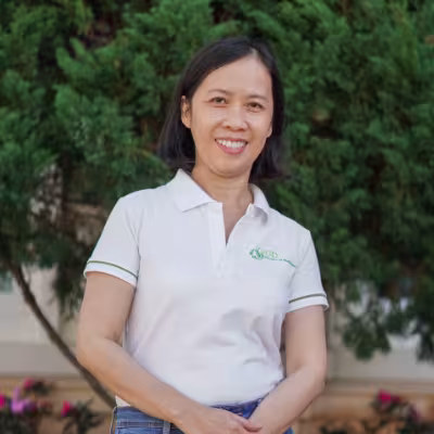 Nhan smiles warmly, wearing a white polo shirt. She stands against a backdrop of lush green foliage.