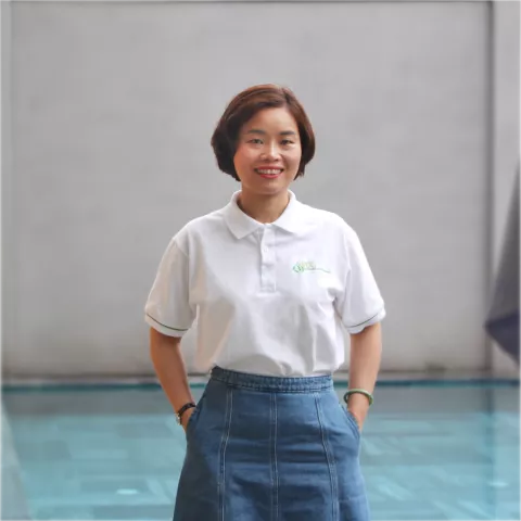 Oanh wearing a white polo shirt and denim skirt stands by a pool. The mood is friendly and casual, set against a plain background.