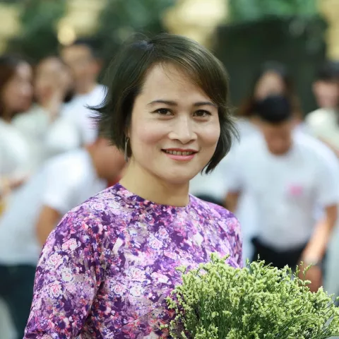 Quyen is wearing purple floral dress smiles warmly, holding a bouquet of delicate white flowers. People in white shirts are blurred in the background.
