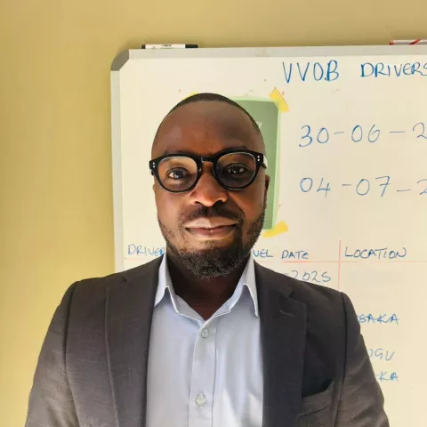 Boyd is wearing a grey suit over a blue shirt and glasses. He is standing against a whiteboard with writing on it.