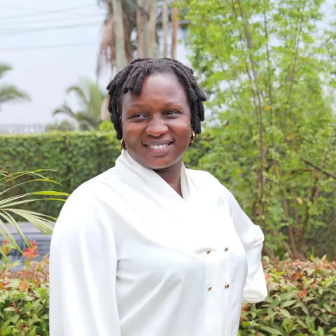 Brenda wears a white shirt and smiles softly. She is standing in front of lush, green plants.