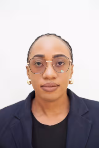 Bridget is wearing round glasses, gold earrings, a black shirt, and a dark blazer, facing forward against a plain white background.