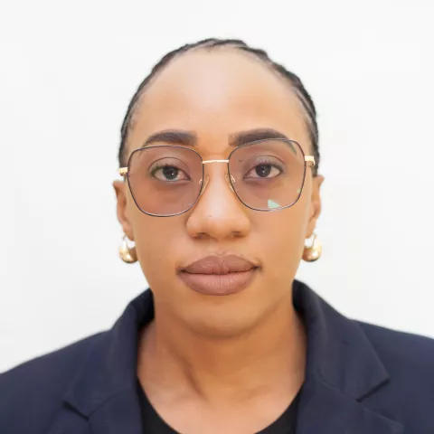 Bridget is wearing round glasses, gold earrings, a black shirt, and a dark blazer, facing forward against a plain white background.