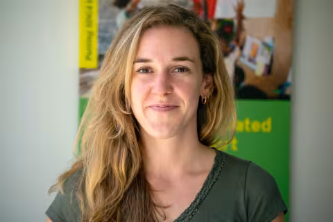 Charlotte smiles softly at the camera. She is wearing a green top. The background features colorful, blurred posters.