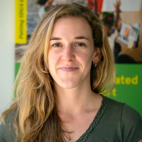Charlotte smiles softly at the camera. She is wearing a green top. The background features colorful, blurred posters.