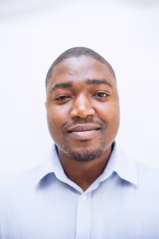 Chipo is wearing a light blue collared shirt and stands against a plain, light background, looking directly at the camera with a neutral expression.