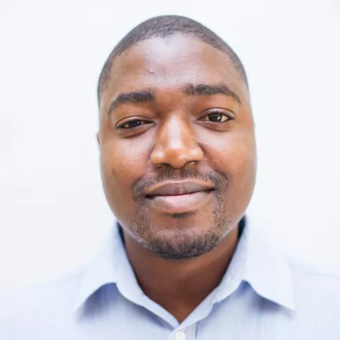 Chipo is wearing a light blue collared shirt and stands against a plain, light background, looking directly at the camera with a neutral expression.