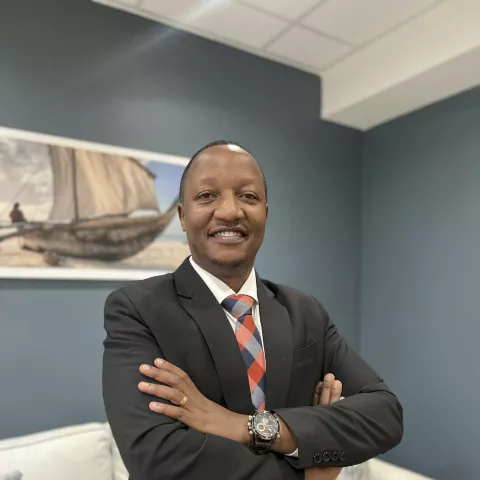 Christopher is wearing a black suit and colorful tie. He stands confidently with arms crossed in an office, a painting of a boat on the wall behind.