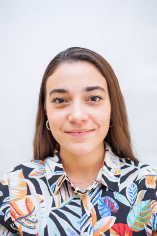 Clara is wearing a colorful shirt and smiles against a white background.