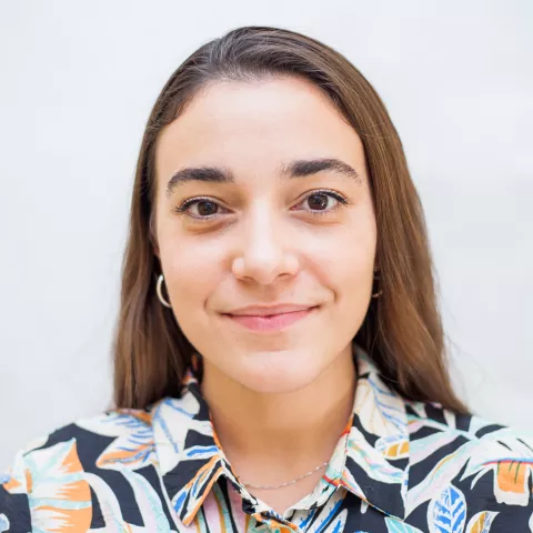 Clara is wearing a colorful shirt and smiles against a white background.