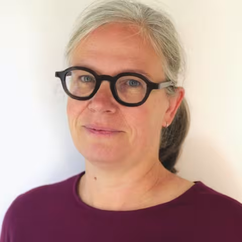 Maud is wearing a red shirt and black glasses. The background is a plain white wall.