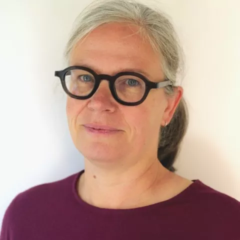 Maud is wearing a red shirt and black glasses. The background is a plain white wall.