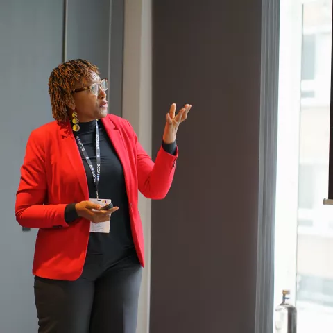 Cleopatra is in red blazer and presents confidently beside a screen displaying bullet points. Her expression is engaged, creating an informative atmosphere.