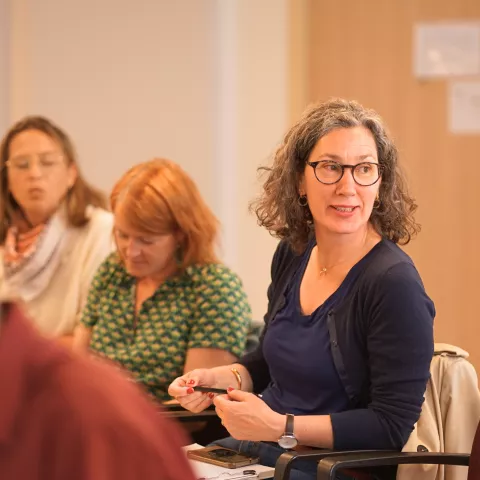 A woman with curly hair and glasses attentively listens in a meeting, surrounded by others. The room conveys a focused and engaged atmosphere.