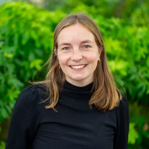 Lisse is wearing a black top and smiles at the camera. The background is green, lush plants.