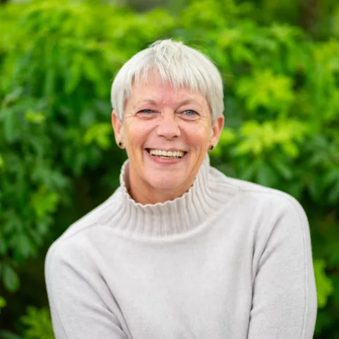 Nancy is wearing a white sweater and smiles. The background is a lush green foliage.