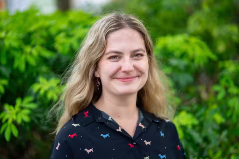 Larissa is wearing a patterned black shirt. She stands in front of lush green foliage.