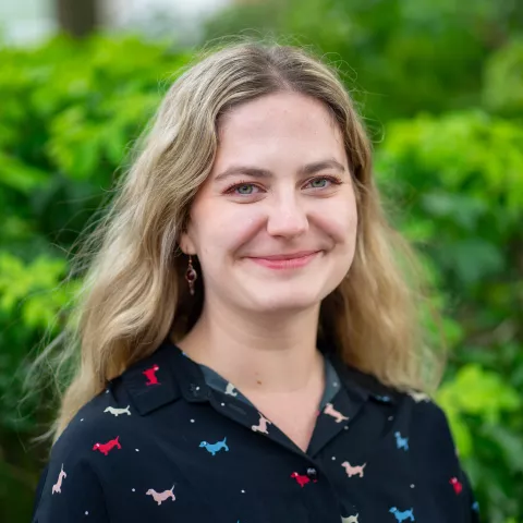 Larissa is wearing a patterned black shirt. She stands in front of lush green foliage.