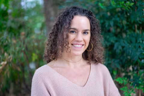 Britt is wearing a light sweater and hoop earrings. She stands smiling in front of lush green foliage.