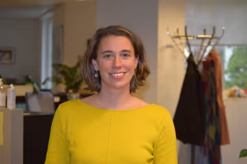 Inge is wearing a yellow top and smiling at the camera. In the background, a modern office is visible.
