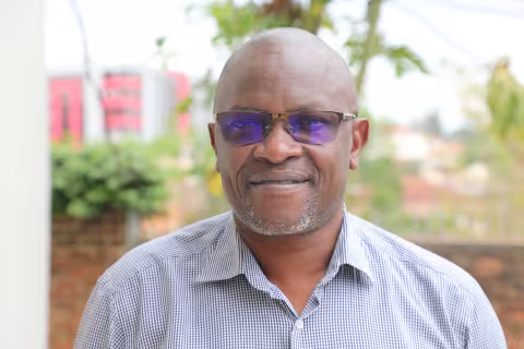 David is wearing a striped shirt and glasses. He is smiling. The background is green plants.