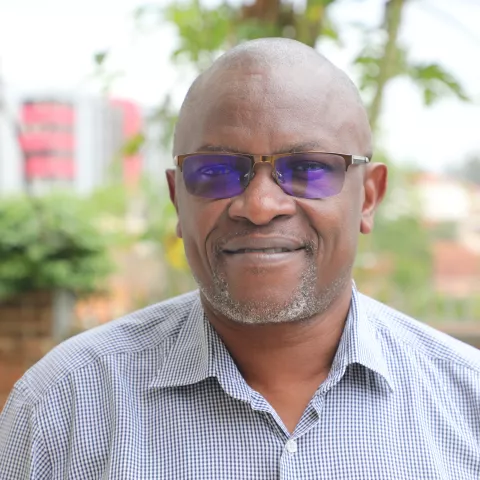 David is wearing a striped shirt and glasses. He is smiling. The background is green plants.