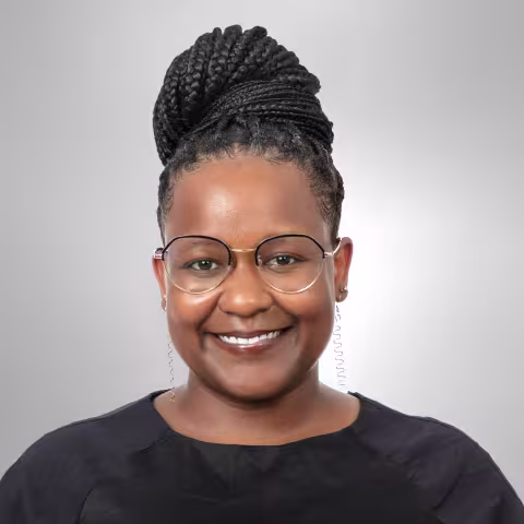 Dorian has braided hair in a bun. She is wearing round glasses and a black top, against a plain gray background.