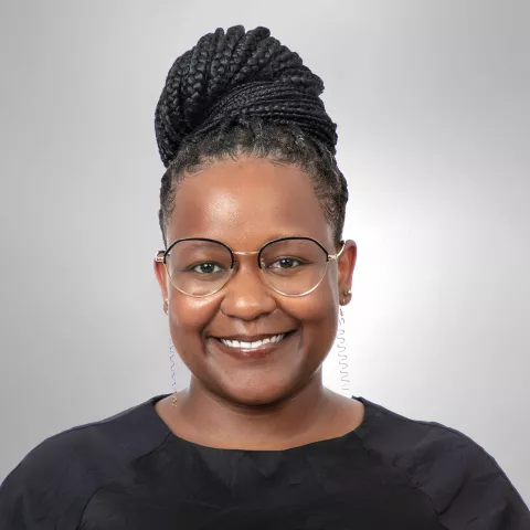 Dorian has braided hair in a bun. She is wearing round glasses and a black top, against a plain gray background.