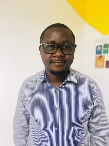 Emmanuel is wearing glasses and a striped shirt. He smiles and stands in front of a white wall with yellow accents.