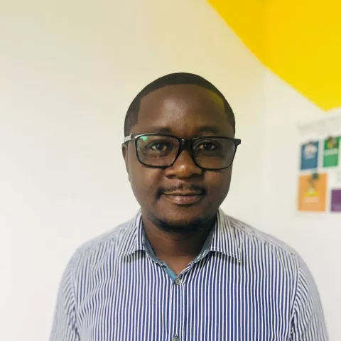 Emmanuel is wearing glasses and a striped shirt. He smiles and stands in front of a white wall with yellow accents.