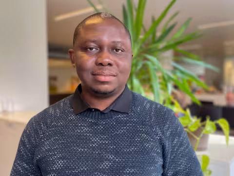 Oyindamola is wearing a dark blue knitted sweater and smiles at the camera. The background is blurred plants.