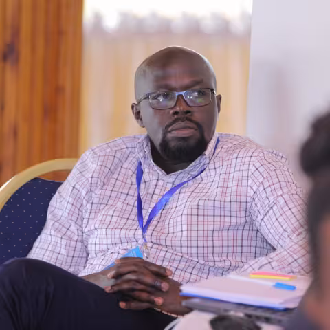 James is wearing glasses and a checked shirt. He sits attentively in a conference room, holding papers. 