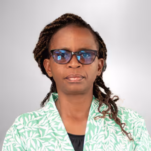 Jane is wearing glasses and stands against a plain gray background. She is wearing a green jacket with a white leaf pattern.