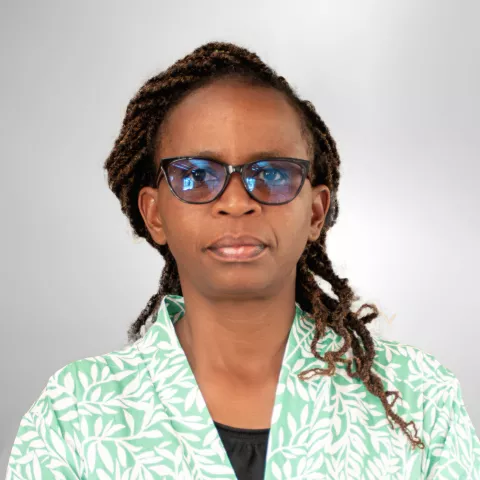 Jane is wearing glasses and stands against a plain gray background. She is wearing a green jacket with a white leaf pattern.