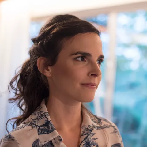 Jasmien is looking away from the camera. She is wearing a patterned shirt. The background is a blurry window.