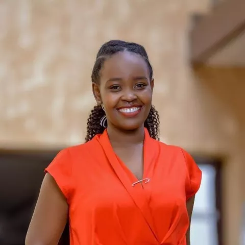 Joanna wears a bright orange shirt. She is smiling softly at the camera. The background is a brown building.