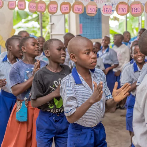 Children in blue uniforms and one in a black t-shirt are clapping in a colorful classroom, decorated with educational posters and paper numbers. They appear joyful and engaged.