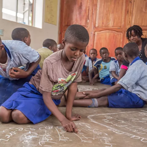 Young learners write on the classroom floor with chalk during a group literacy activity.