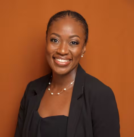 Kakula smiles warmly. She is wearing a black shirt and black blazer and white pearl necklace. She stands in front of a brown backdrop.