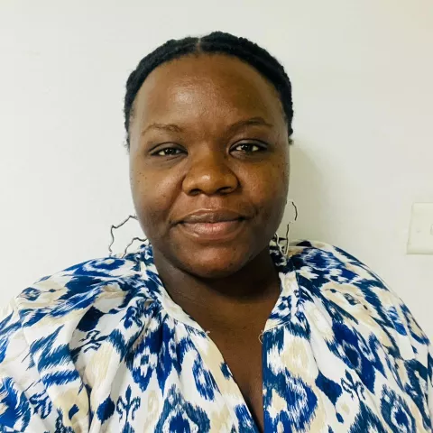 Kalumba is wearing silver earrings and a blue and white patterned top. She is smiling against a white wall. 
