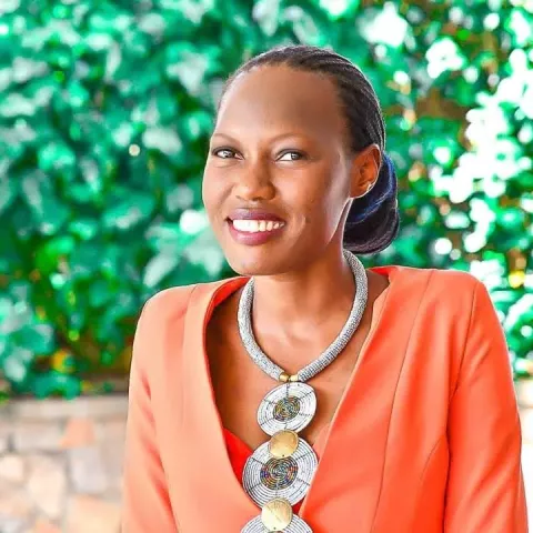 Brendah is wearing an orange jacket with a patterned necklace stands against a leafy green background.