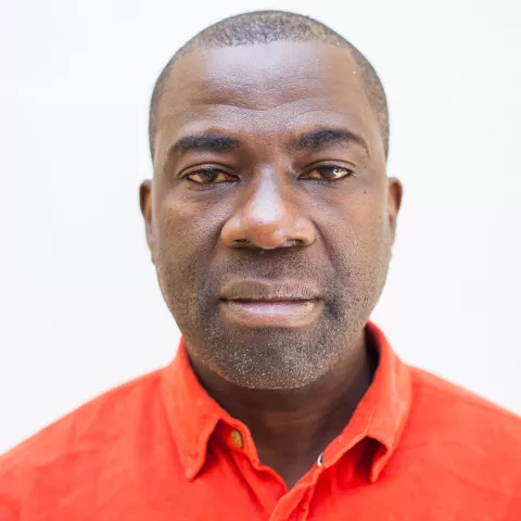 Kennedy is in an orange shirt against a light background, displaying a calm, neutral expression.