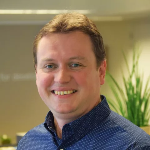 Koenraad is smiling while wearing a blue shirt. The background is a blurred office.