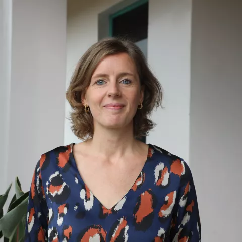 Loran stands smiling in front of a white wall. She wears a patterned blouse and small earrings