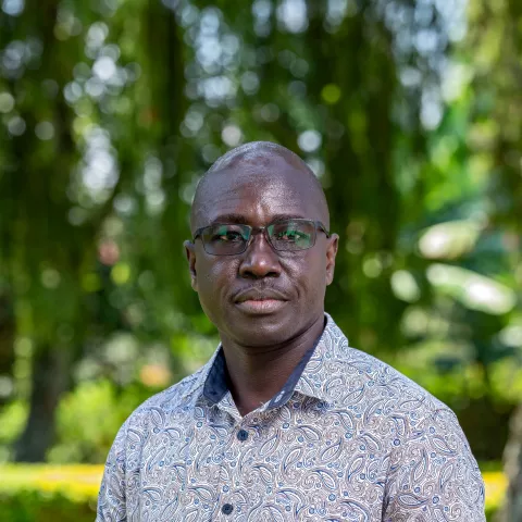 Louis is wearing glasses and a paisley-patterned shirt stands outdoors, looking serious. The background is lush with greenery.