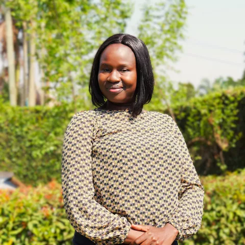 Loy Lyn woman stands outside, smiling softly. She's wearing a patterned blouse. The background features lush green foliage and trees.