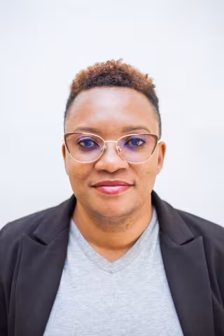 Maimbolwa is wearing glasses and smiles warmly. She is wear a grey shirt and a black blazer, conveying a friendly, professional look. The background is plain white.