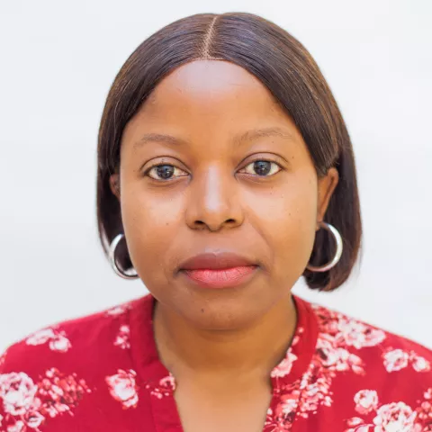 Mercy has hoop earrings and a straight expression. She is wearing a red floral blouse. The background is plain white.
